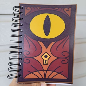 May include: A spiral bound notebook with a colorful cover featuring a large yellow eye with a black pupil and a keyhole design. The cover has a red, orange, and brown color scheme.