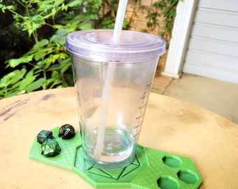 Può includere: Un sottobicchiere verde stampato in 3D con sei incavi esagonali per i dadi. Un bicchiere di plastica trasparente con una cannuccia è appoggiato sul sottobicchiere. Tre dadi verdi e dorati sono sul sottobicchiere.