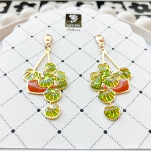 May include: Gold-toned dangle earrings featuring a plant design. Each earring has a gold triangle top with a red-orange pot and green leaves. The earrings are on a white card with a black dotted pattern.
