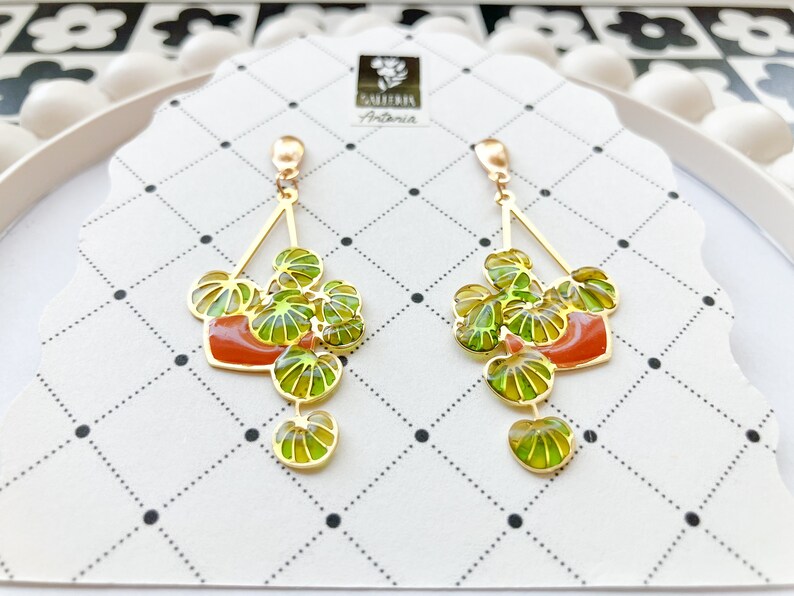 May include: Gold-toned dangle earrings featuring a plant design. Each earring has a gold triangle top with a red-orange pot and green leaves. The earrings are on a white card with a black dotted pattern.