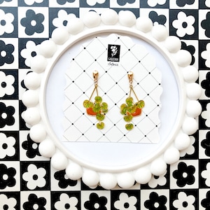 May include: A pair of gold-toned earrings featuring a plant design. Each earring has a small orange pot with green leaves and hanging vines. The earrings are displayed on a white card with a black and white checkered background.