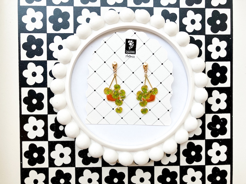 May include: A pair of gold-toned earrings featuring a plant design. Each earring has a small orange pot with green leaves and hanging vines. The earrings are displayed on a white card with a black and white checkered background.