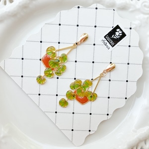May include: Gold-toned earrings featuring a plant design with green and yellow leaves and a red pot. The earrings are on a white card with a black dotted grid pattern. The card has a logo that says "Galleria Antonia".