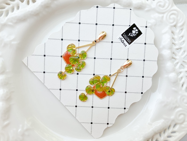 May include: Gold-toned earrings featuring a plant design with green and yellow leaves and a red pot. The earrings are on a white card with a black dotted grid pattern. The card has a logo that says "Galleria Antonia".