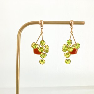 May include: A pair of dangle earrings featuring a plant design. Each earring has a red pot with green and yellow leaves. The earrings are suspended from a gold-colored metal hook.