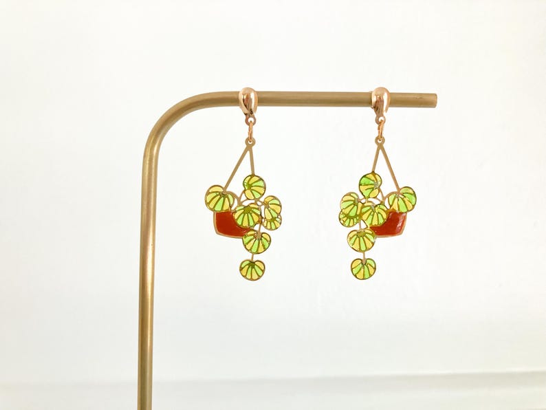 May include: A pair of dangle earrings featuring a plant design. Each earring has a red pot with green and yellow leaves. The earrings are suspended from a gold-colored metal hook.