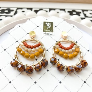 May include: A pair of gold-toned hoop earrings with multiple layers of beads in shades of orange, cream, and yellow. The bottom of the earrings feature brown, faceted beads. The earrings are displayed on a patterned card with the text "Galleria Antania".
