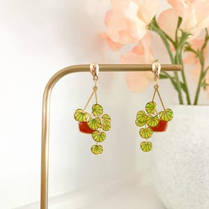 May include: Gold-toned dangle earrings featuring a plant design. Each earring has a red pot with green and yellow leaves. The earrings are displayed on a gold earring stand, with a vase of flowers in the background.