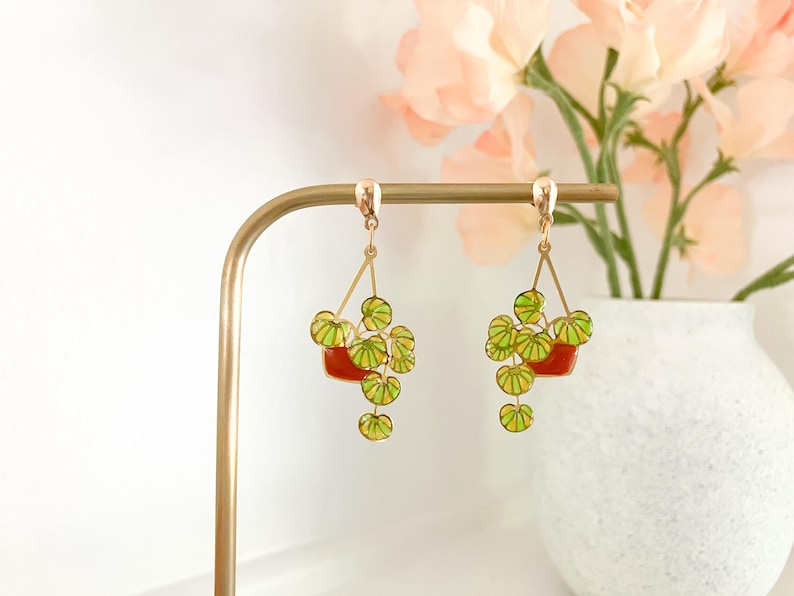 May include: Gold-toned dangle earrings featuring a plant design. Each earring has a red pot with green and yellow leaves. The earrings are displayed on a gold earring stand, with a vase of flowers in the background.