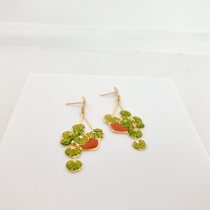 May include: Gold-toned dangle earrings featuring a plant design. Each earring has a gold frame with a red-orange pot and green and yellow leaves. The earrings are displayed on a white surface.