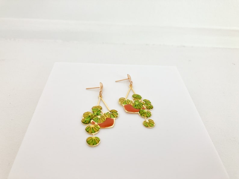 May include: Gold-toned dangle earrings featuring a plant design. Each earring has a gold frame with a red-orange pot and green and yellow leaves. The earrings are displayed on a white surface.