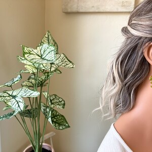 May include: Gold-toned dangle earrings with a floral design featuring green and orange accents. The earrings are worn by a person with gray and blonde hair. A potted plant with large green and white leaves is in the background.