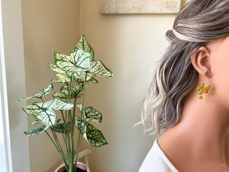 May include: Gold-toned dangle earrings with a floral design featuring green and orange accents. The earrings are worn by a person with gray and blonde hair. A potted plant with large green and white leaves is in the background.