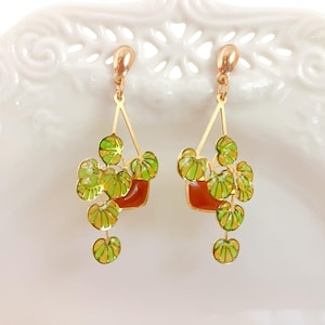 May include: Gold-tone dangle earrings featuring a plant design. Each earring has a small, square, reddish-brown pot with green and yellow leaves. The leaves hang down from the pot. The earrings are set against a white background.
