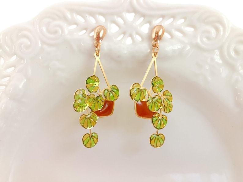 May include: Gold-tone dangle earrings featuring a plant design. Each earring has a small, square, reddish-brown pot with green and yellow leaves. The leaves hang down from the pot. The earrings are set against a white background.