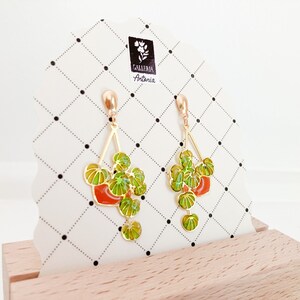 May include: Gold-tone earrings featuring a plant design with green and yellow leaves and a red pot. The earrings are displayed on a white card with a black and white grid pattern and the text "GALLERIA Antonia".