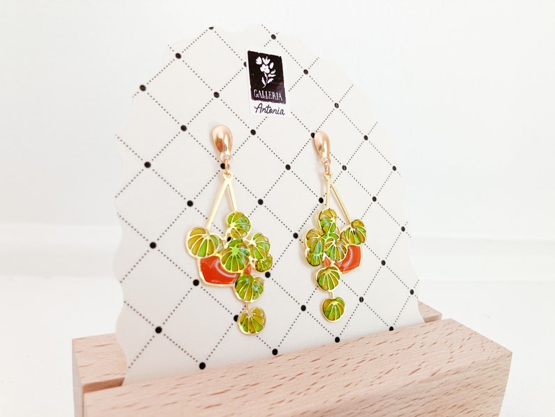 May include: Gold-tone earrings featuring a plant design with green and yellow leaves and a red pot. The earrings are displayed on a white card with a black and white grid pattern and the text "GALLERIA Antonia".