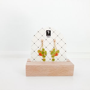 May include: Gold-colored dangle earrings featuring a plant-in-pot design. The earrings have a triangular shape with a red pot and green leaves. The earrings are displayed on a white card with a black grid pattern and a wooden base.