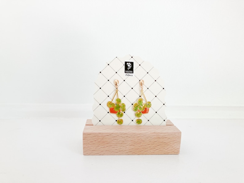 May include: Gold-colored dangle earrings featuring a plant-in-pot design. The earrings have a triangular shape with a red pot and green leaves. The earrings are displayed on a white card with a black grid pattern and a wooden base.