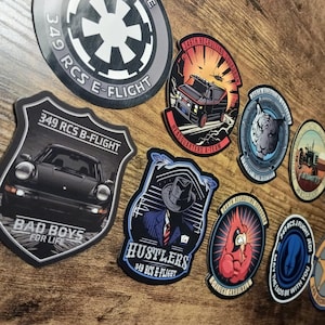 May include: A collection of various stickers with different designs and text. The stickers feature images of a car, a tiger, a cardinal, and a star. Text includes "349 RCS E-FLIGHT", "BAD BOYS FOR LIFE", and "HUSTLERS".
