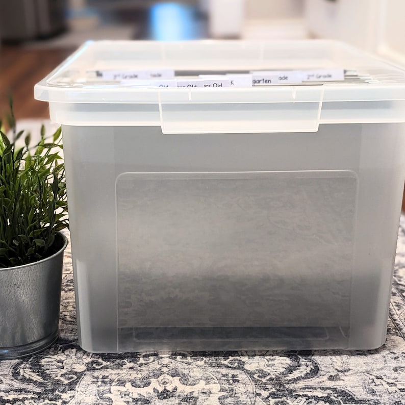 May include: A clear plastic storage bin with a gray label on the front. The label has the words "Kindergarten", "1st Grade", "2nd Grade", and "3rd Grade" written on it.