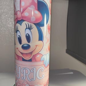 Pode incluir: Um copo cil&iacute;ndrico com o gr&aacute;fico de um rato de desenho animado com um la&ccedil;o rosa de bolinhas, sobre um fundo rosa claro com pequenos cora&ccedil;&otilde;es. A parte inferior do copo exibe o nome YRIC em letras brancas com contorno.