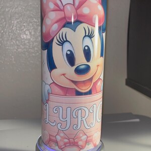 Pode incluir: Um copo cil&iacute;ndrico com um gr&aacute;fico da Minnie Mouse usando um la&ccedil;o rosa de bolinhas. A personagem est&aacute; posicionada acima de uma faixa rosa claro contendo o nome LYRIC em letras mai&uacute;sculas brancas. O design inclui pequenos cora&ccedil;&otilde;es.