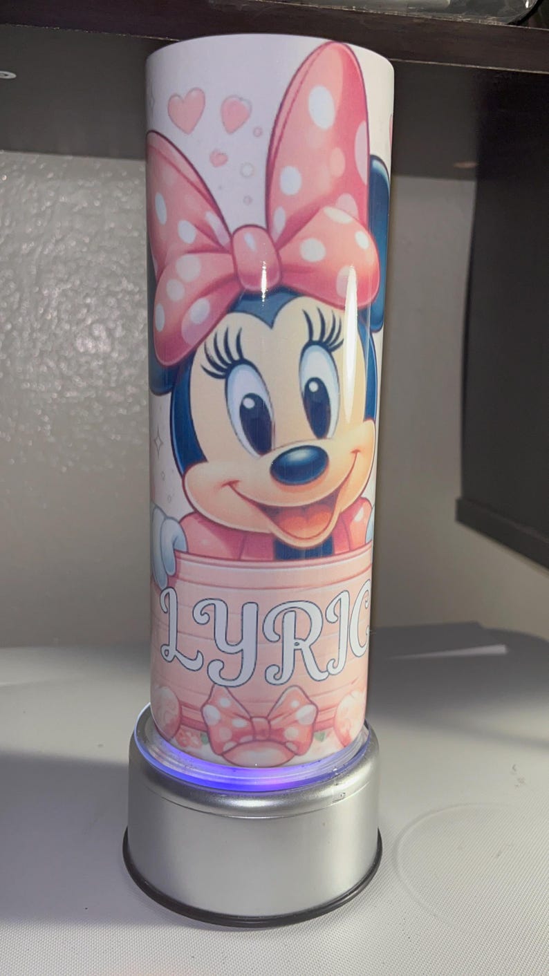 Pode incluir: Um copo cil&iacute;ndrico com um gr&aacute;fico da Minnie Mouse usando um la&ccedil;o rosa de bolinhas. A personagem est&aacute; posicionada acima de uma faixa rosa claro contendo o nome LYRIC em letras mai&uacute;sculas brancas. O design inclui pequenos cora&ccedil;&otilde;es.