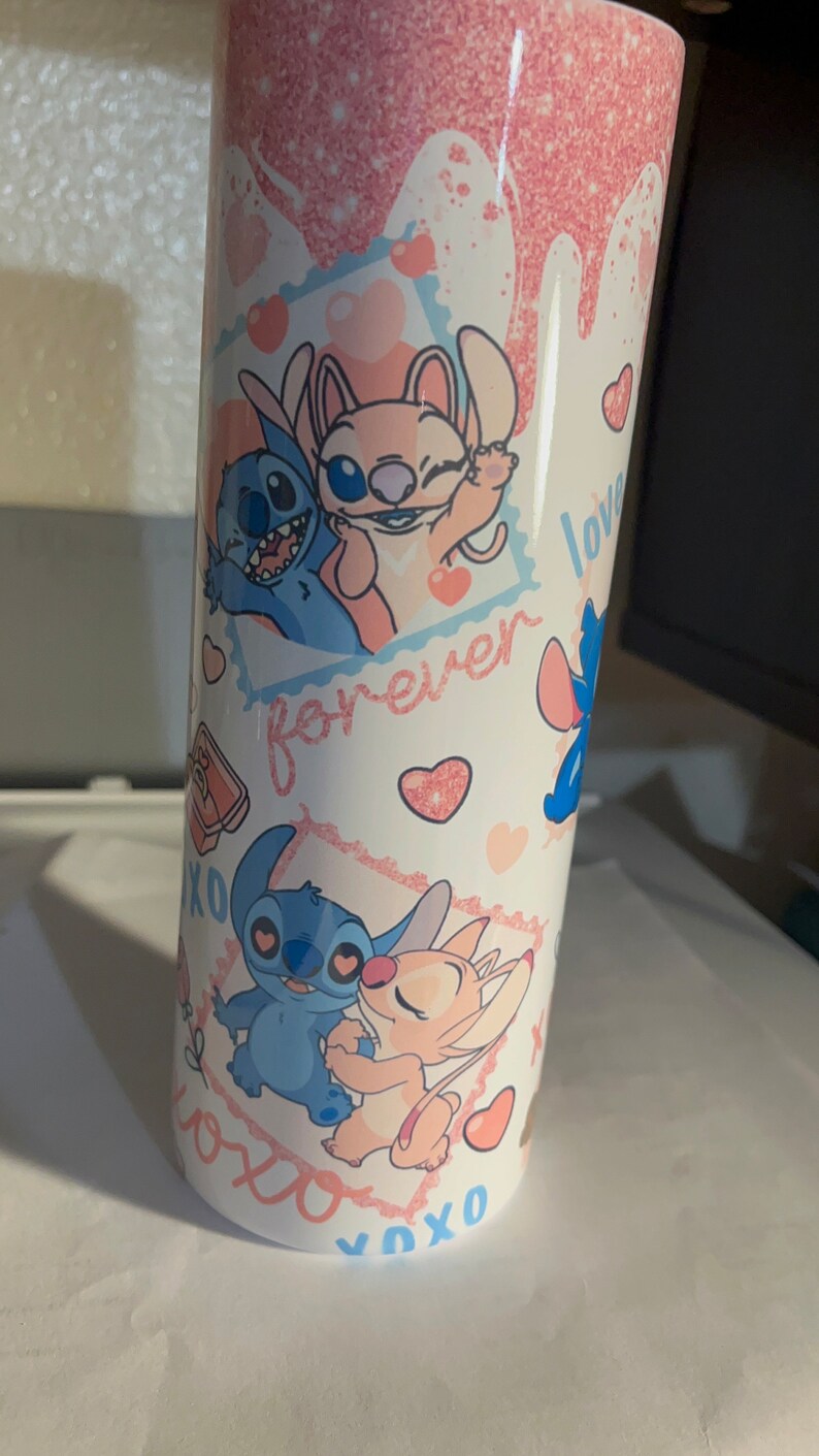 May include: A cylindrical tumbler with a white base featuring a pink glitter drip effect at the top. The design includes illustrations of blue and pink alien characters, red hearts, and the text "forever" and "XOXO" printed in a cursive pink font.