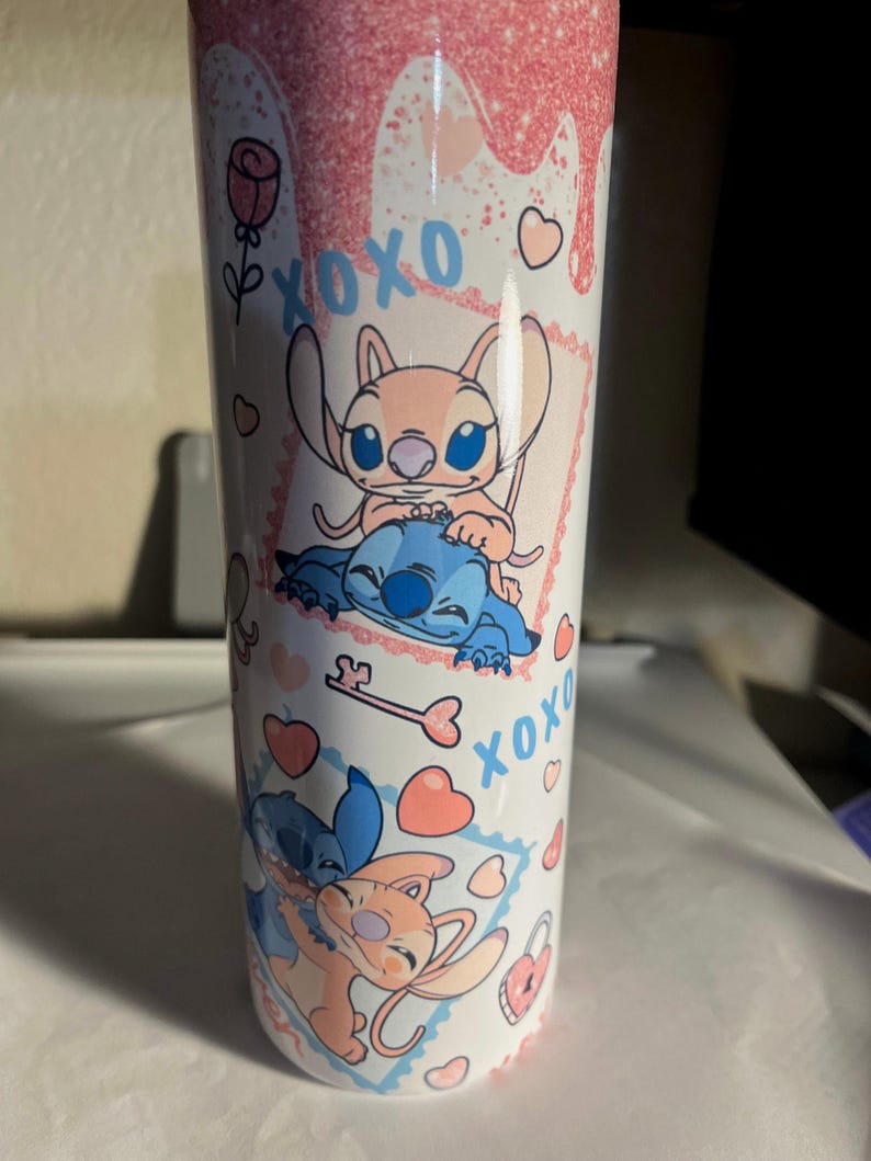 May include: A white cylindrical tumbler featuring a pink glitter drip design at the top. The surface is decorated with blue and pink cartoon characters, the text XOXO, pink hearts, a rose, and a key. The characters are shown in two separate frames.
