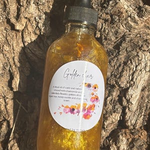 May include: A golden glass bottle with a black dropper top and a white label that reads "Golden Elixir". The label also includes a description of the product, which is a ritual oil of calm and radiance infused with chamomile and calendula flowers, golden shimmer, tiger's eye, honey suckle and chamomile scent. The bottle is sitting on a brown, textured surface.