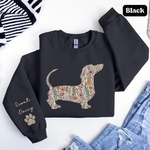 May include: Black sweatshirt featuring a floral dachshund design. The dog silhouette is filled with a pattern of pink, orange, and green flowers. The left sleeve has the text "Scout Daisy" and a paw print.