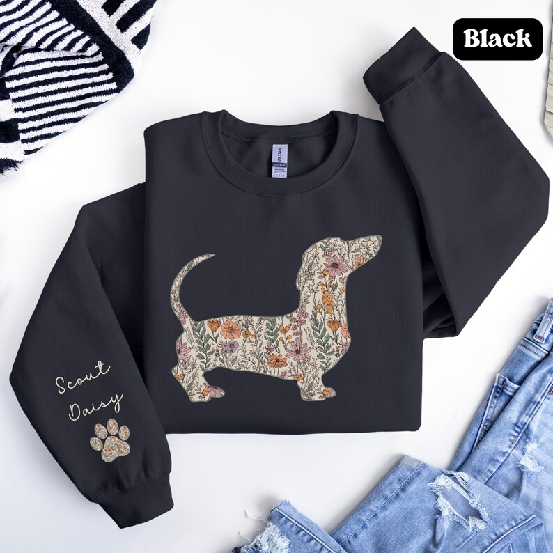 May include: Black sweatshirt featuring a floral dachshund design. The dog silhouette is filled with a pattern of pink, orange, and green flowers. The left sleeve has the text "Scout Daisy" and a paw print.