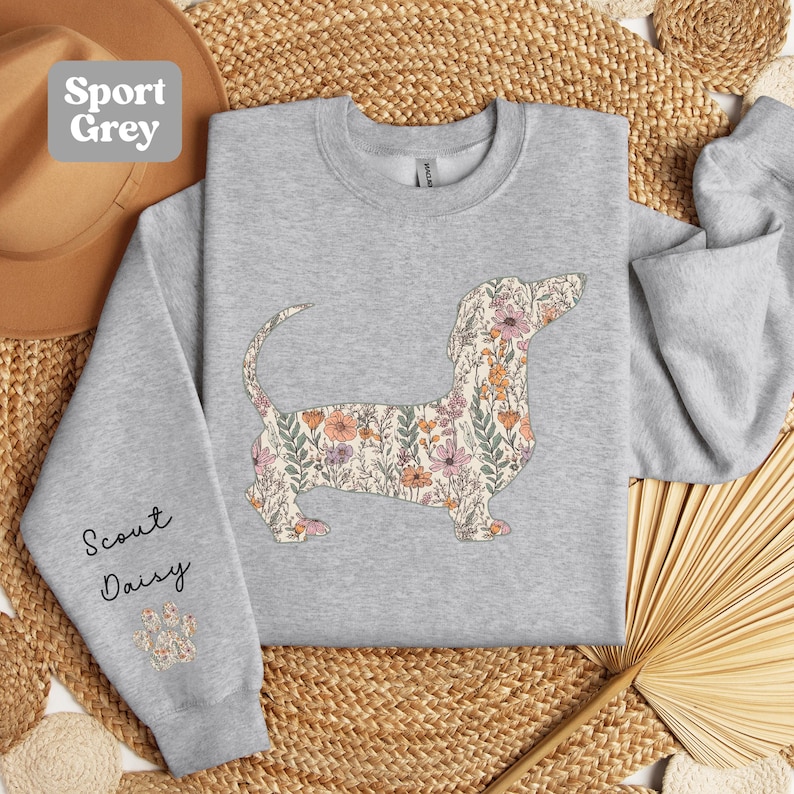 May include: Sport Grey sweatshirt featuring a floral dachshund design. The dog silhouette is filled with a pattern of pink, orange, and green flowers. The left sleeve has the text "Scout Daisy" and a paw print.