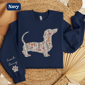 May include: Navy blue sweatshirt featuring a floral dachshund design. The dog silhouette is filled with a pattern of pink, orange, and green flowers. The left sleeve has the text "Scout Daisy" and a paw print.