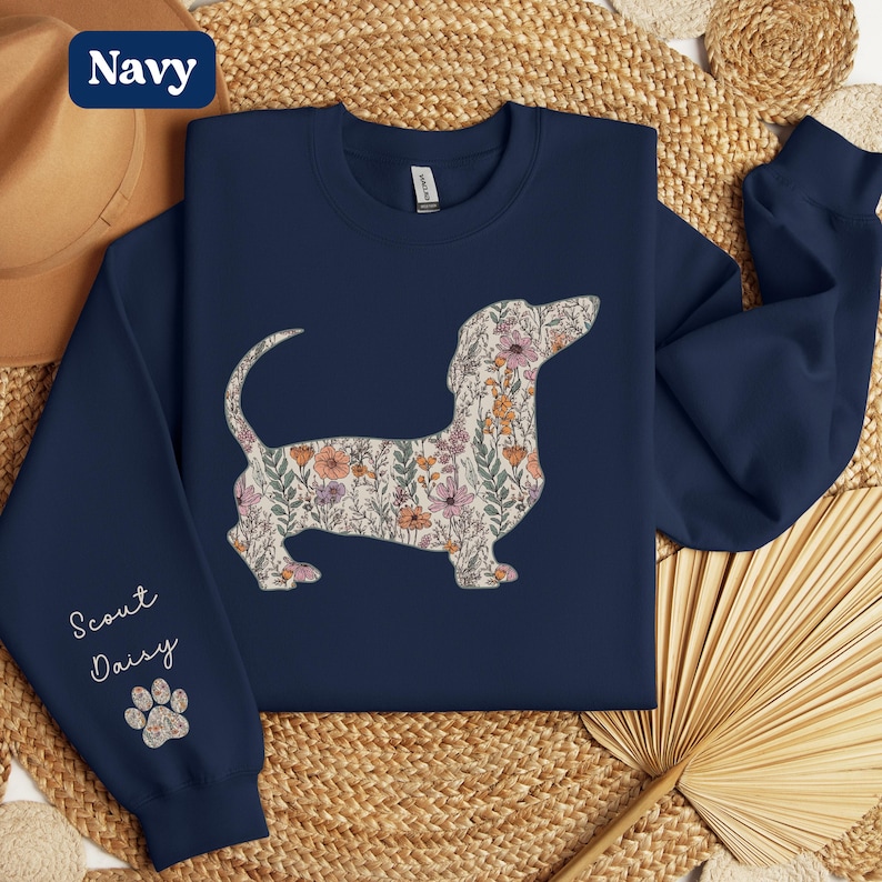 May include: Navy blue sweatshirt featuring a floral dachshund design. The dog silhouette is filled with a pattern of pink, orange, and green flowers. The left sleeve has the text "Scout Daisy" and a paw print.