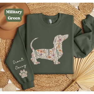 May include: Military green sweatshirt featuring a floral dachshund design. The dog silhouette is filled with a pattern of pink, orange, and green flowers. The sleeve reads "Scout Daisy" with a paw print.