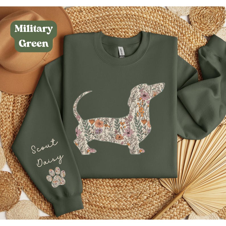 May include: Military green sweatshirt featuring a floral dachshund design. The dog silhouette is filled with a pattern of pink, orange, and green flowers. The sleeve reads "Scout Daisy" with a paw print.