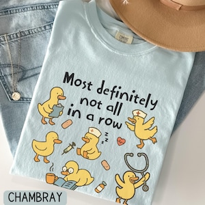 May include: Light blue t-shirt with the text "Most definitely not all in a row" and cartoon ducks in various medical roles. The shirt is laid on top of blue jeans and a tan hat.