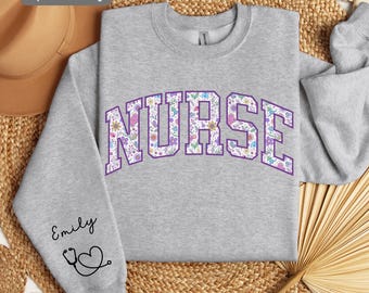 May include: A gray sweatshirt with the word "NURSE" printed in white letters outlined in purple. The letters are filled with a colorful floral pattern. The sweatshirt has a stethoscope and heart graphic on the sleeve with the name "Emily" written above it.