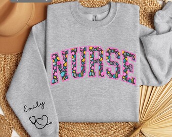 May include: A gray sweatshirt with the word "NURSE" printed in pink letters with a floral pattern. The word is in an arch shape. The sweatshirt has a small stethoscope and heart design on the left sleeve with the name "Emily" written above it.
