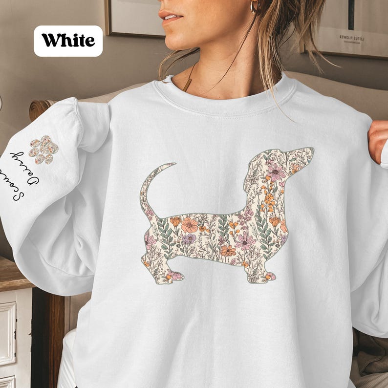 May include: White sweatshirt featuring a floral dachshund design. The dog silhouette is filled with a pattern of flowers in shades of pink, orange, and green. The left sleeve has the text "Scout Daisy" and a paw print design. The word "White" is in a white box.