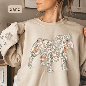 Op de afbeelding: Zandkleurige sweatshirt met een bloemen bulldog ontwerp. Het bulldog silhouet is gevuld met een patroon van roze, oranje en groene bloemen en bladeren. De linkermouw heeft een pootafdruk ontwerp en de woorden "Sco Daisy" verticaal gedrukt.