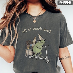 May include: A charcoal grey t-shirt featuring a graphic of a green frog wearing a pointed hat and riding a white scooter. The frog carries a crate filled with maths supplies, including an abacus and ruler. The text reads, off to teach math.