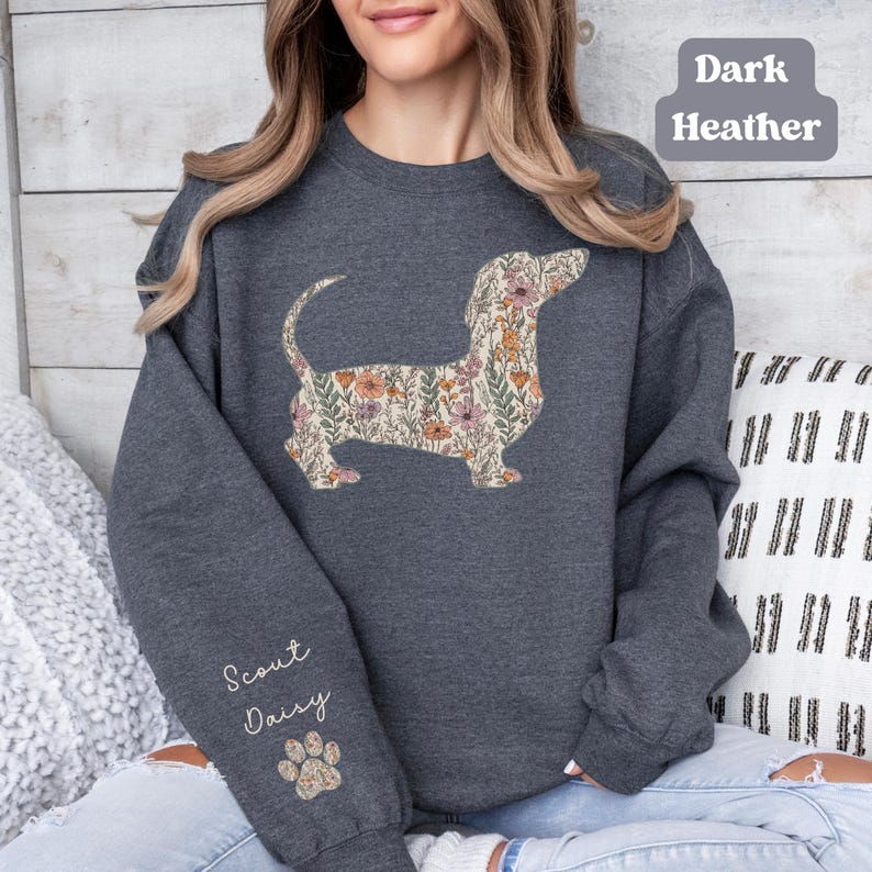 May include: Dark heather gray sweatshirt featuring a floral dachshund design. The dog silhouette is filled with a pattern of flowers in shades of pink, orange, and green. The sleeve has the text "Scout Daisy" and a paw print.