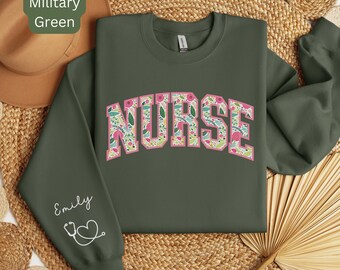 May include: A military green sweatshirt with the word "NURSE" in pink letters with a floral pattern. The word "NURSE" is in a collegiate font. The left sleeve has the name "Emily" and a stethoscope with a heart.
