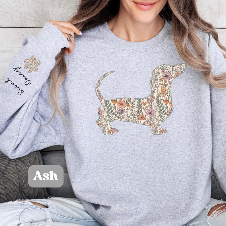 May include: Ash-colored sweatshirt featuring a floral dachshund design. The dachshund is filled with a pattern of flowers in shades of pink, orange, and green. The left sleeve has the words "Scout Dairy" and a paw print. The word "Ash" is printed on the bottom left.