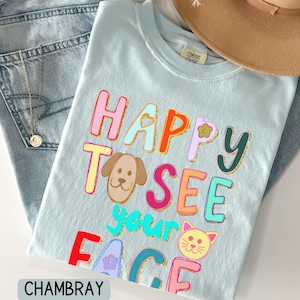 May include: A light blue t-shirt featuring the colorful text HAPPY TO SEE your FACE with a cartoon dog and cat illustration. The letters are outlined in gold glitter. The shirt has a Comfort Colors label at the inner neckline.