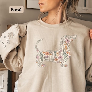 May include: Sand-colored sweatshirt featuring a floral dachshund design. The dog silhouette is filled with pink, orange, and green flowers. The left sleeve has the names "Scout" and "Daisy" printed vertically, with a floral paw print above.