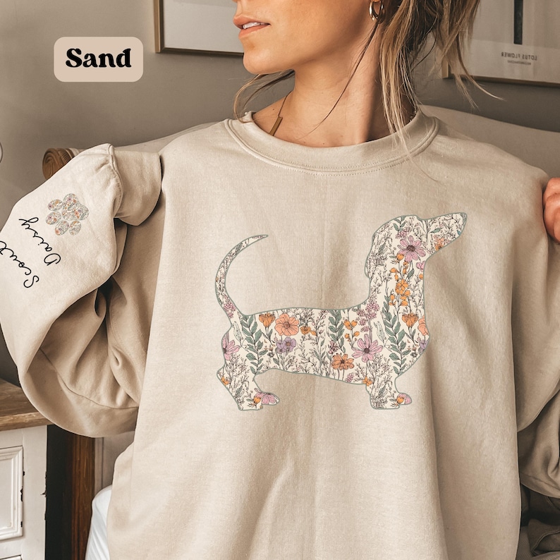 May include: Sand-colored sweatshirt featuring a floral dachshund design. The dog silhouette is filled with pink, orange, and green flowers. The left sleeve has the names "Scout" and "Daisy" printed vertically, with a floral paw print above.